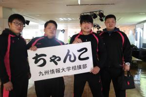 九州情報大学相撲部の男子学生4人が、ちゃんこ鍋の看板を持ってこちらに笑いかけている様子