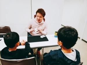 日本経済大学の男子学生が、子供たち２人を前に、机の上でマジックする様子