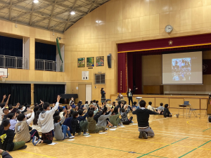 体育館に太宰府西小学校の小学生が集まり、ステージ上のスクリーンに百済初等学校の小学生が映されている。お別れの時間で、ステージ前カメラに向かって手を振る太宰府西小の生徒たちの写真。