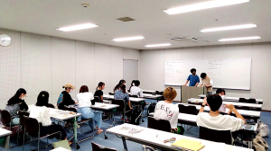 ホワイトボードの前で司会と初期の学生が話しており、そのほかの学生は考え込んでいる様子