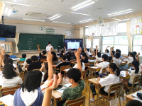 教室を後方から見た様子。黒板横のディスプレイに映された奈良市側発表のクイズに、国分小学校の生徒が手を挙げて回答しています。